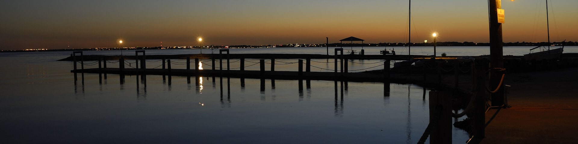 A calm waterfront at dusk with a pier extending into the water, street lamps, and distant city lights.