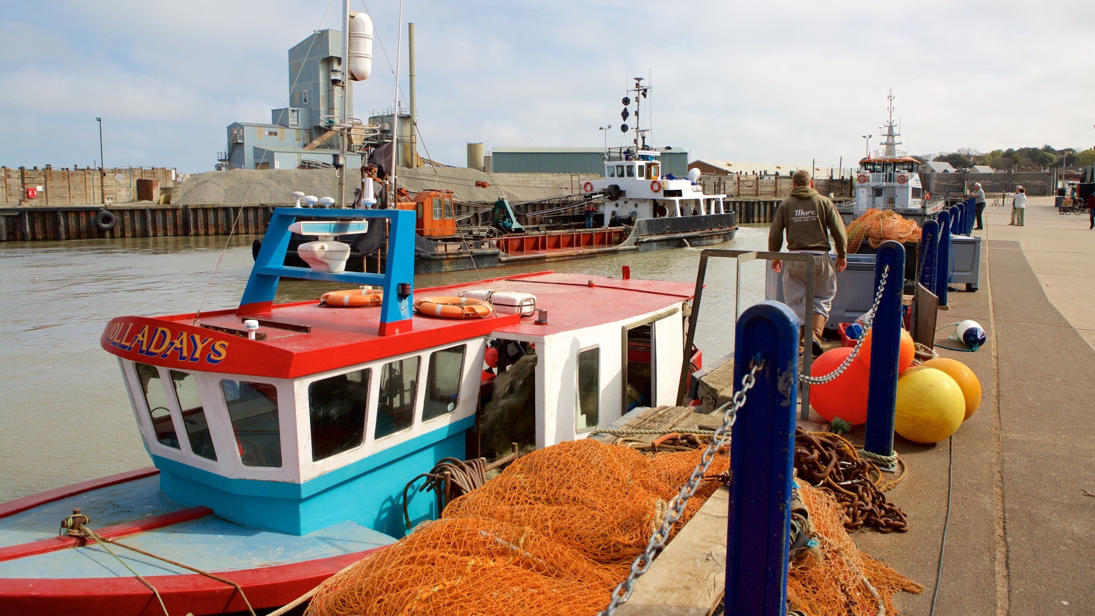 Um cais com um barco vermelho e azul chamado "HOLLADAYS" e redes de pesca laranja.