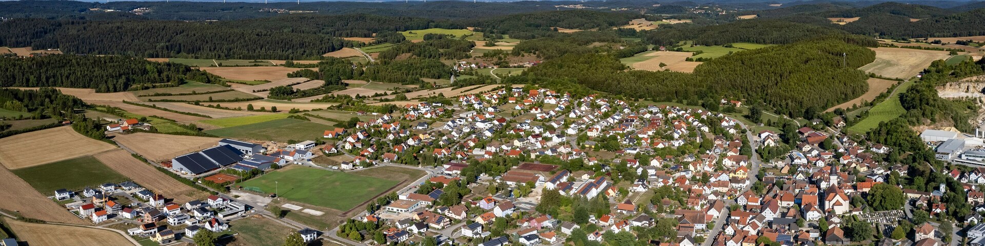 Vista aérea de un pueblo rodeado de campos y bosques, con aerogeneradores a lo lejos.