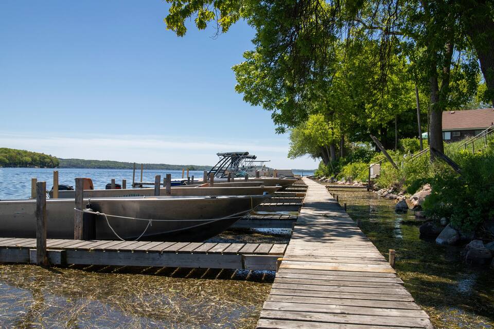 A wooden dock with boats docked along a calm lake, surrounded by trees and a clear sky.