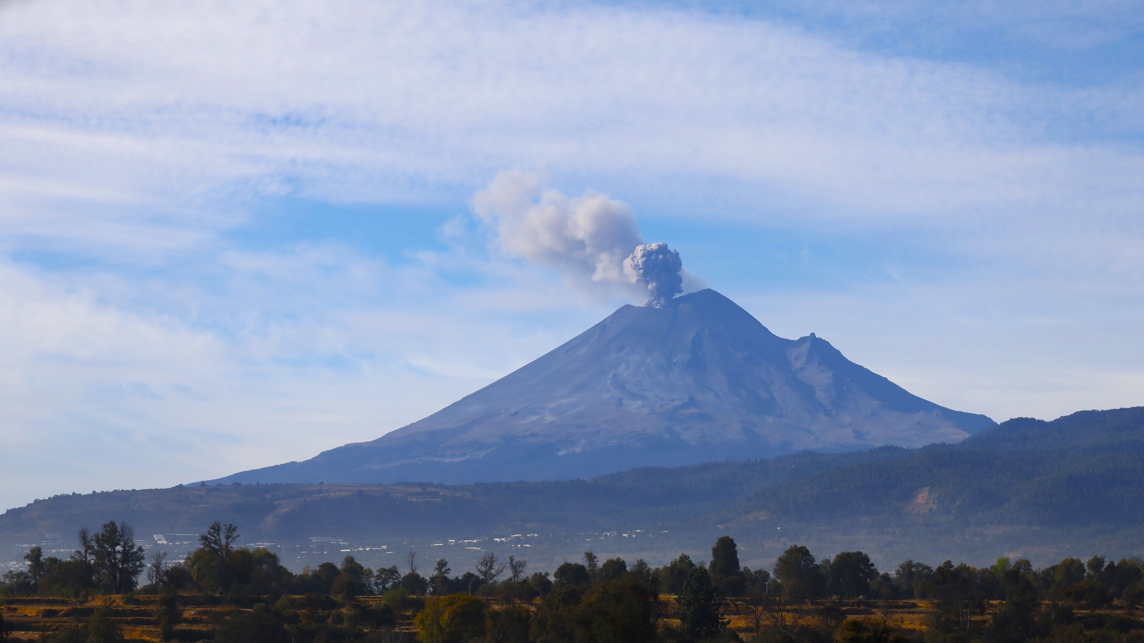 A volcano with smoke rising, surrounded by a forested landscape and distant buildings.