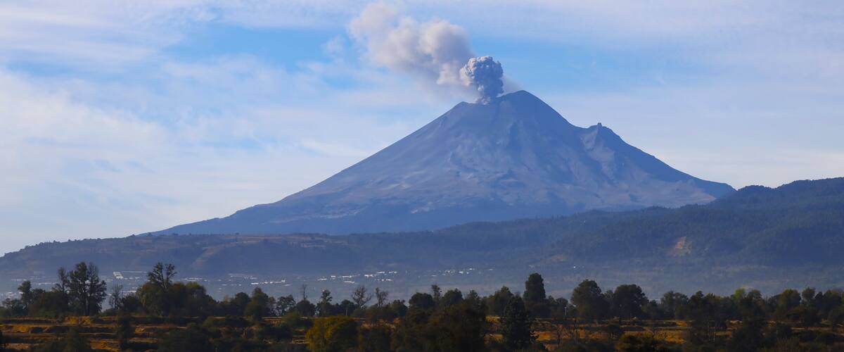 A volcano with smoke rising, surrounded by a forested landscape and distant buildings.