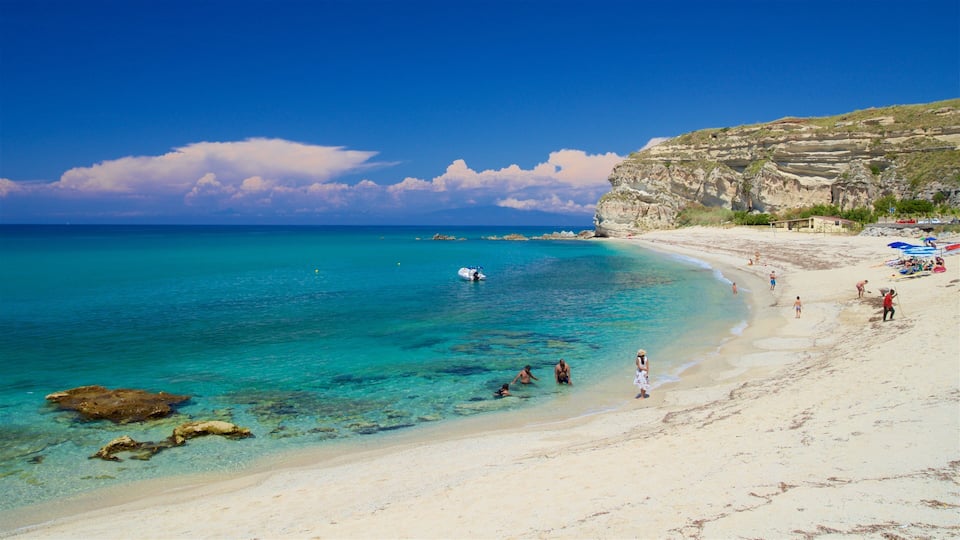 A beach with clear turquoise water, rocky outcrops, and a sandy shore with people and umbrellas.