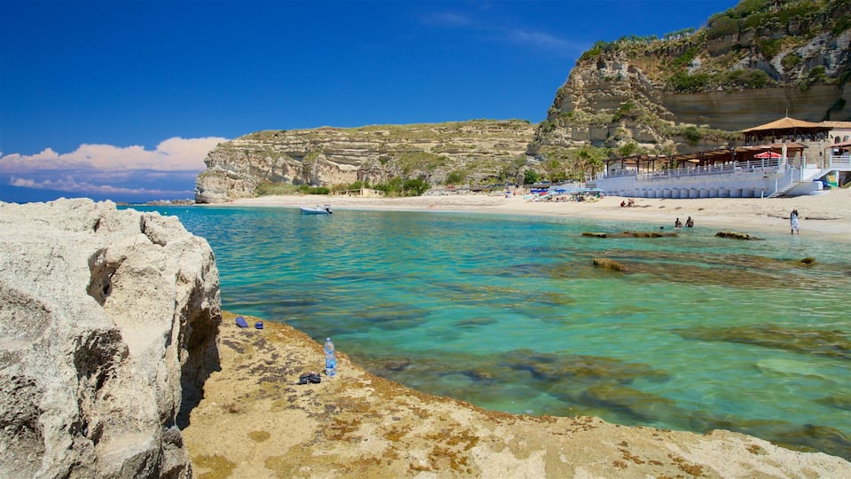 A coastal landscape with clear turquoise waters, rocky cliffs, and a sandy beach with umbrellas.