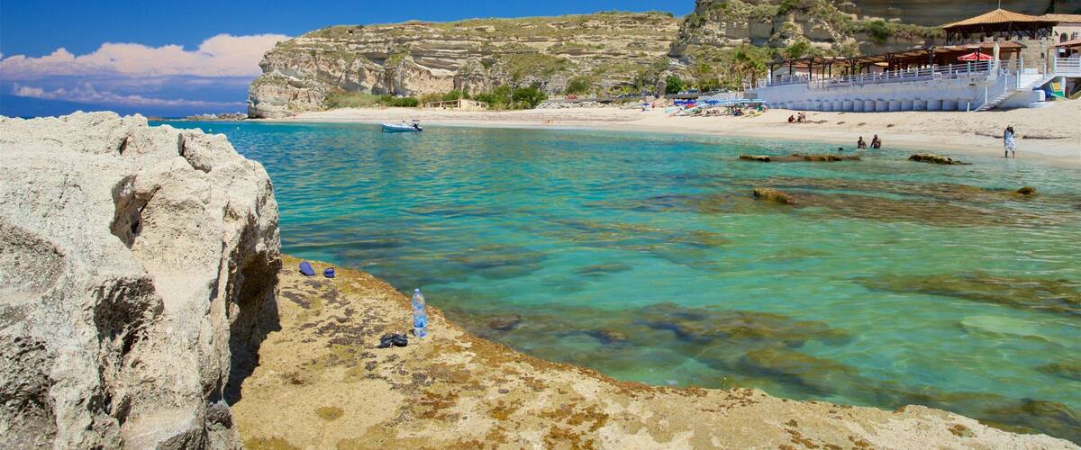 A coastal landscape with clear turquoise waters, rocky cliffs, and a sandy beach with umbrellas.
