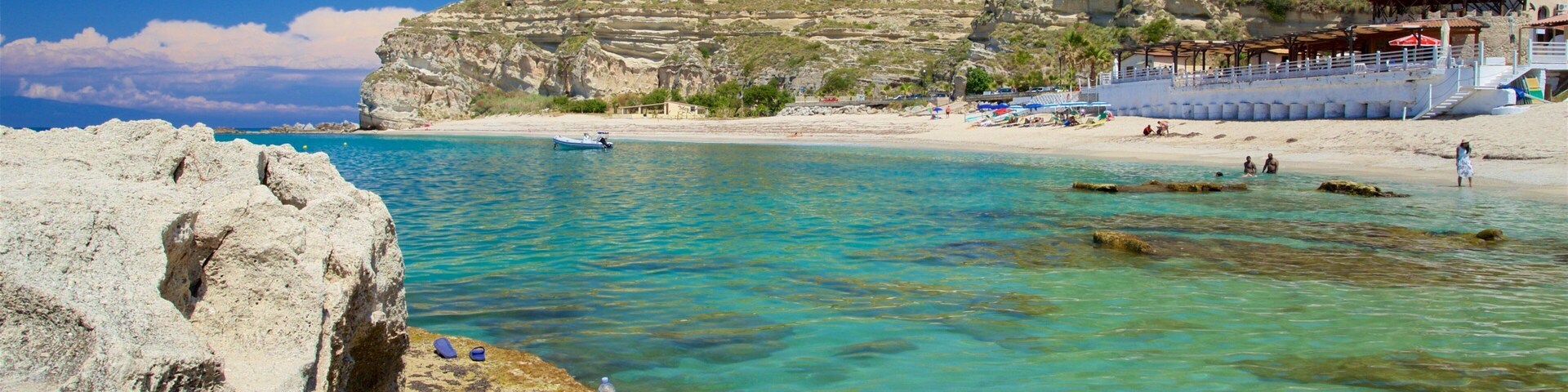A coastal landscape with clear turquoise waters, rocky cliffs, and a sandy beach with umbrellas.