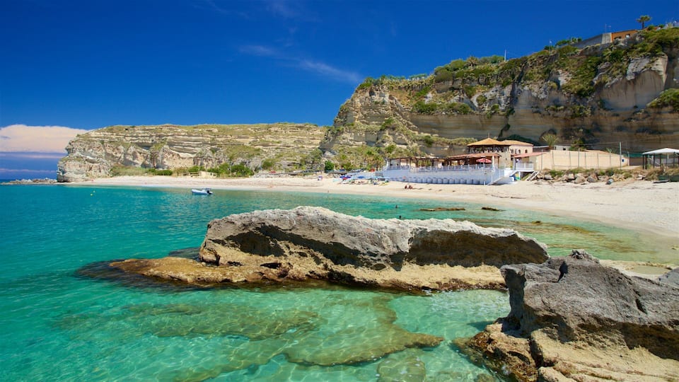 A coastal beach with clear turquoise waters, rocky formations, and a cliff with vegetation.