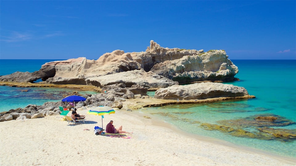 A beach with clear turquoise water, rocky formations, and people relaxing under colorful umbrellas.
