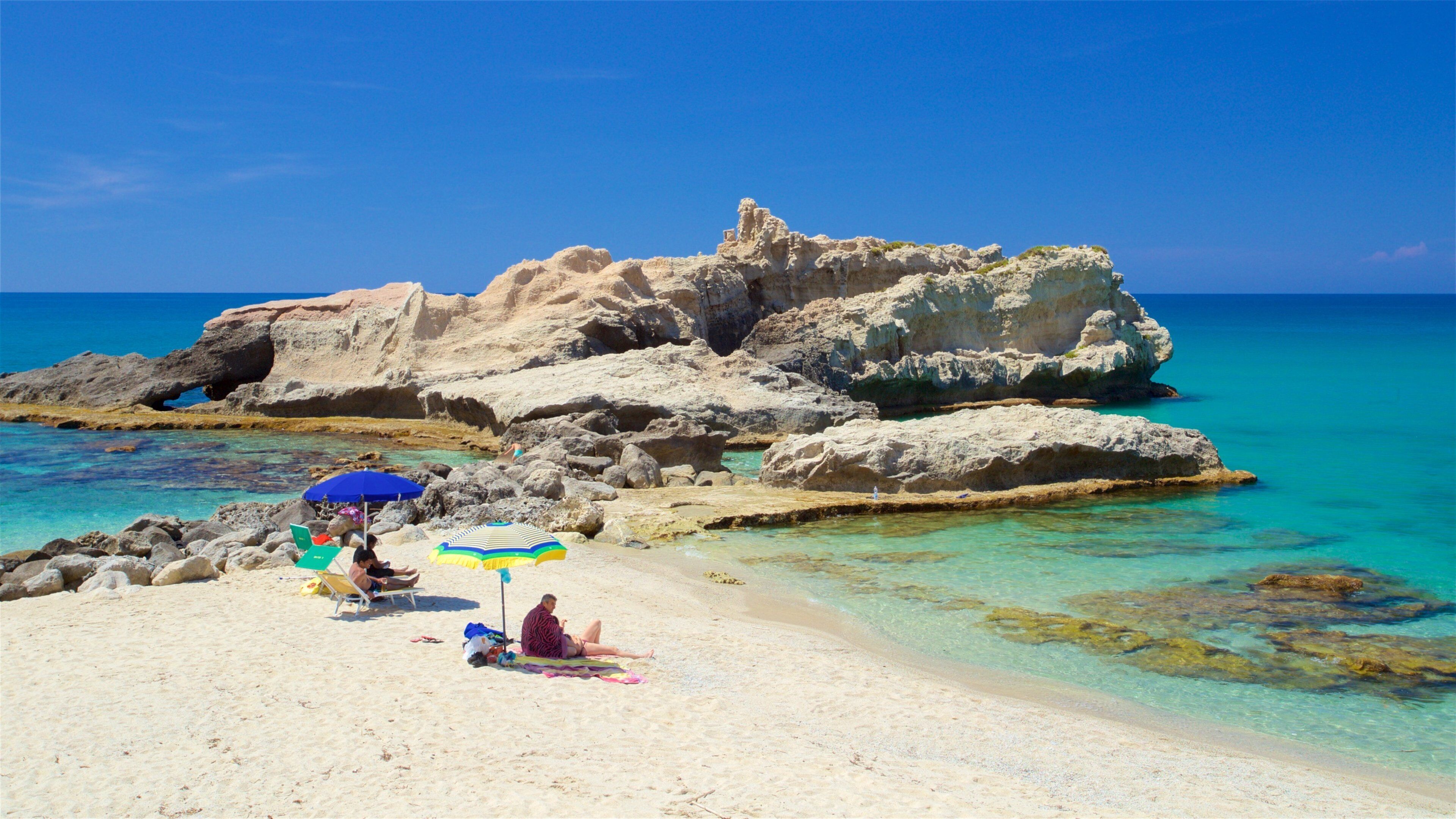 A beach with clear turquoise water, rocky formations, and people relaxing under colorful umbrellas.