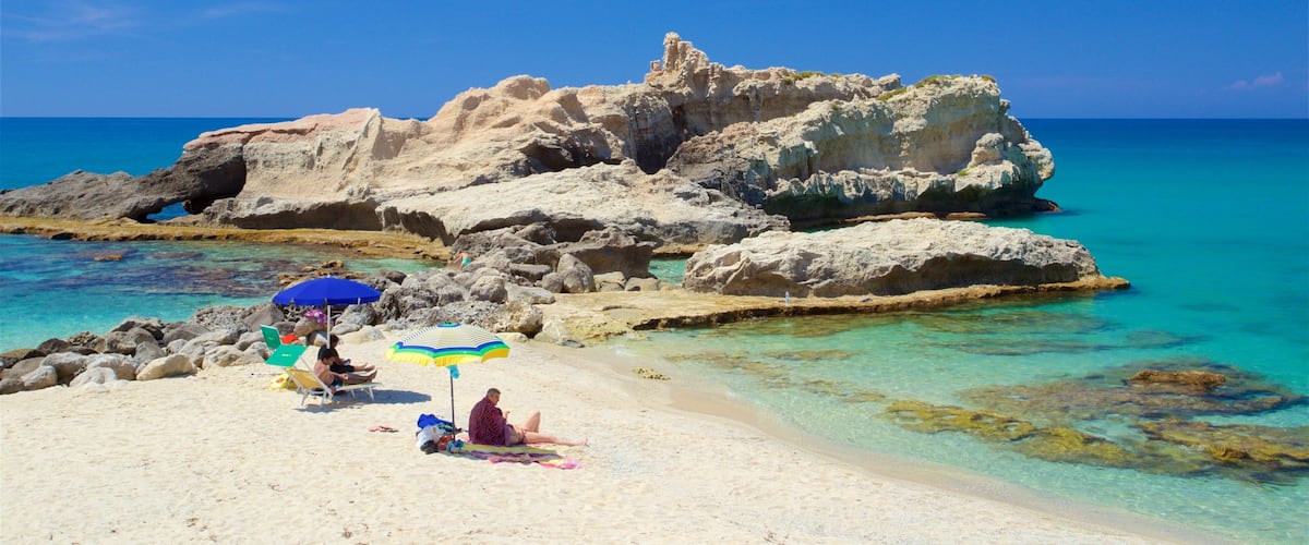 A beach with clear turquoise water, rocky formations, and people relaxing under colorful umbrellas.