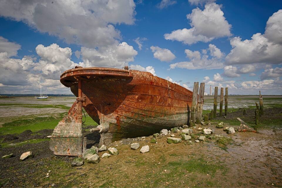 A rusted shipwreck on a beach with a clear sky and a distant sailboat.