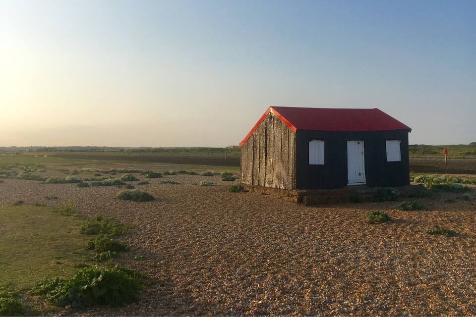 Un petit bâtiment avec un toit rouge et des portes blanches, entouré de gravier et d’une végétation clairsemée, situé dans un paysage rural.