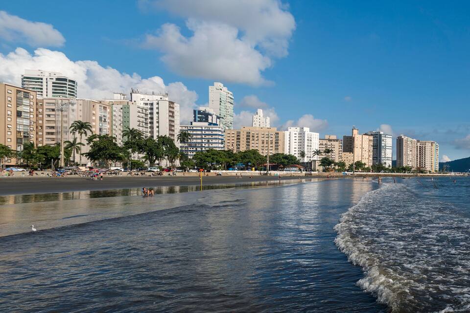 A coastal cityscape with a beach, palm trees, and high-rise buildings under a partly cloudy sky.