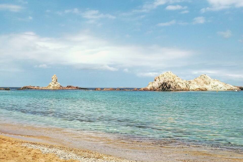 A beach with clear turquoise water and rocky formations in the distance.