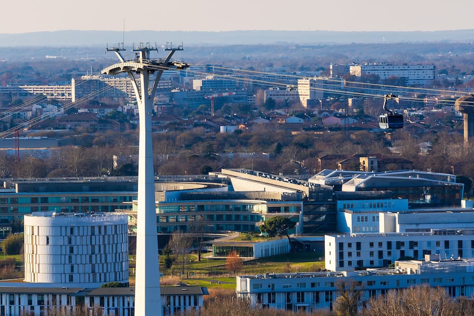 Una torre blanca alta amb un sistema de telefèric, envoltada d'edificis i arbres.