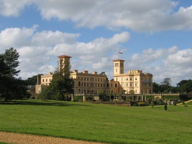 A large, historic building with a central tower and symmetrical wings, surrounded by a well-maintained lawn and trees.