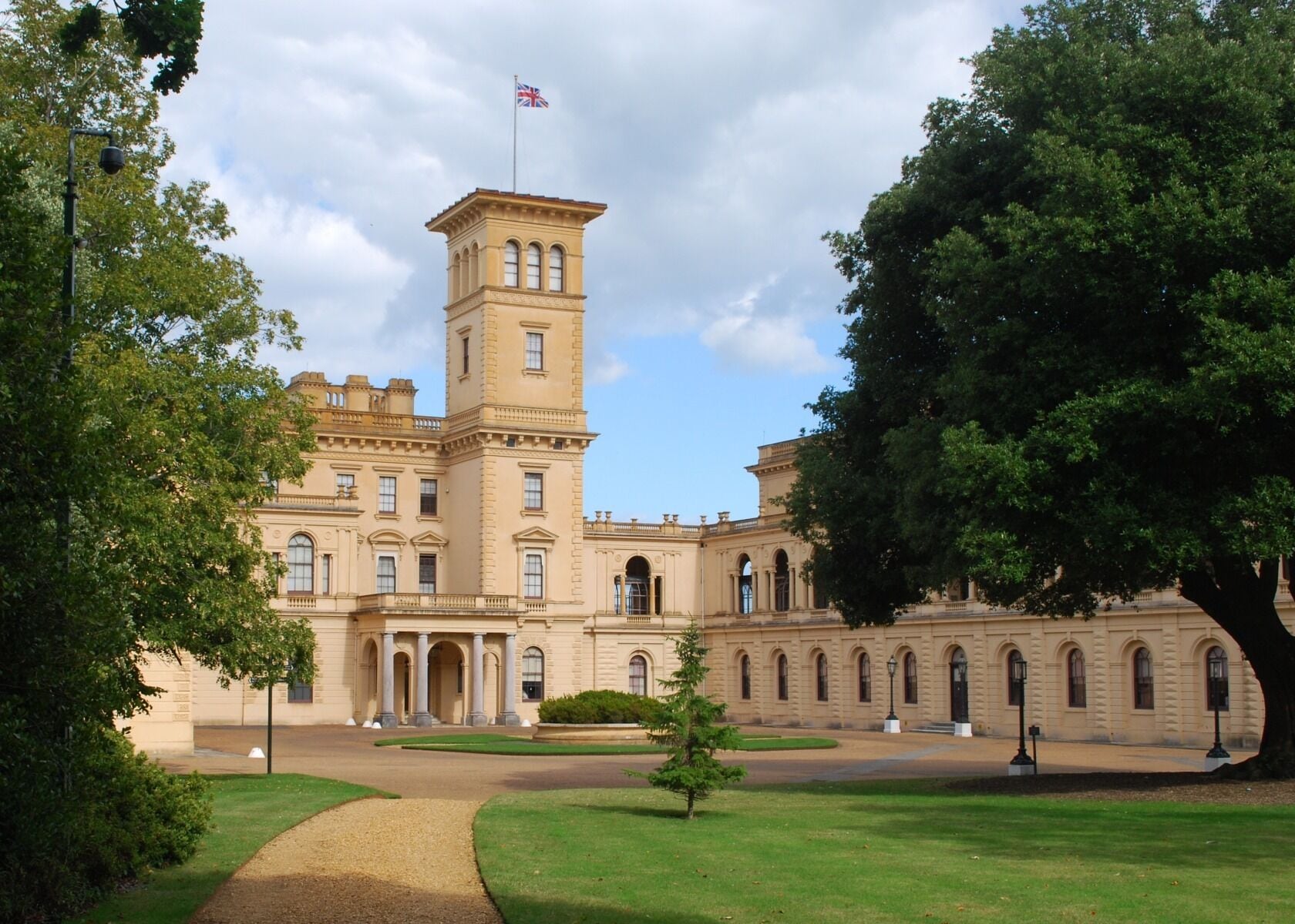 A large, historic building with a prominent tower and flag, surrounded by lush greenery and a well-maintained lawn.