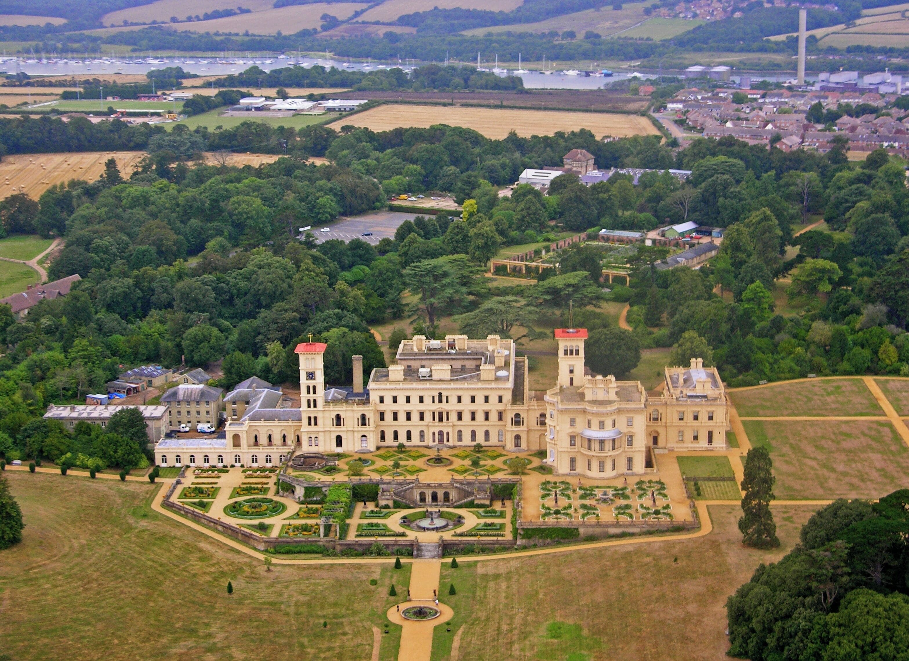 An aerial view of a large estate with a central building featuring two towers, surrounded by manicured gardens and fields.