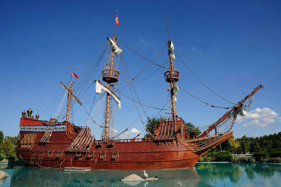 A large wooden ship with multiple masts and flags, docked in a calm body of water with a clear blue sky in the background.