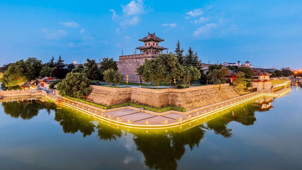 A traditional Chinese pagoda on a hill overlooking a calm river, surrounded by trees and modern buildings in the background.