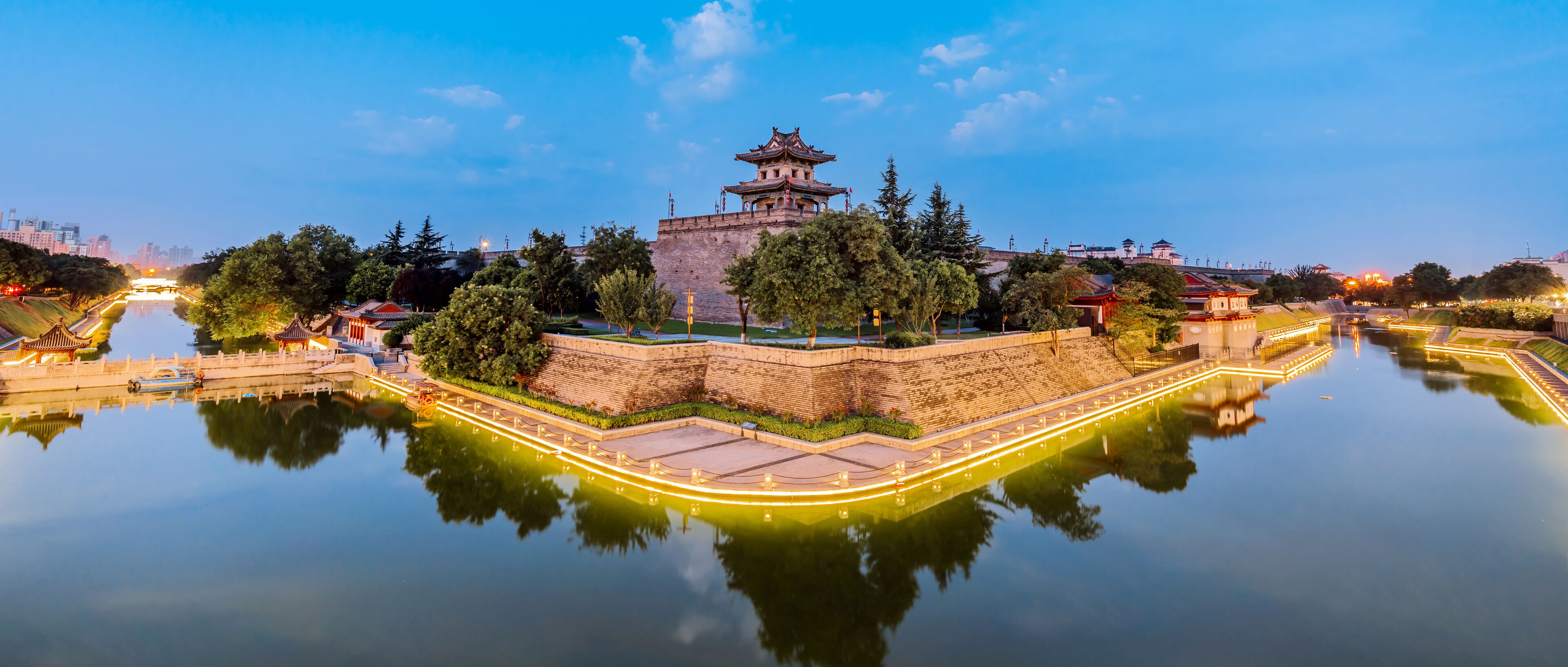 A traditional Chinese pagoda on a hill overlooking a calm river, surrounded by trees and modern buildings in the background.