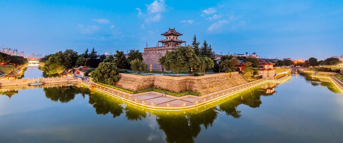 A traditional Chinese pagoda on a hill overlooking a calm river, surrounded by trees and modern buildings in the background.