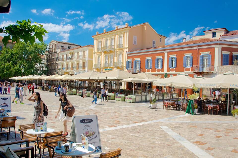 Un espace café animé en plein air, doté de tables et de chaises, ainsi que de parasols, et entouré de bâtiments historiques aux toits recouverts de tuiles rouges.
