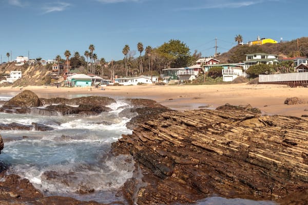 Une côte rocheuse où les vagues s’écrasent sur le rivage, une plage de sable et des maisons à flanc de colline.