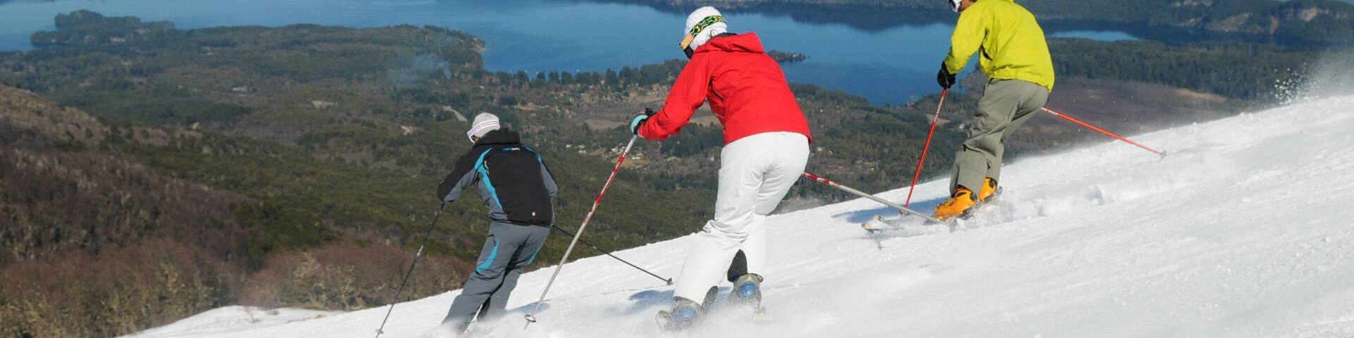 Three skiers on a snowy slope with a lake and mountains in the background.