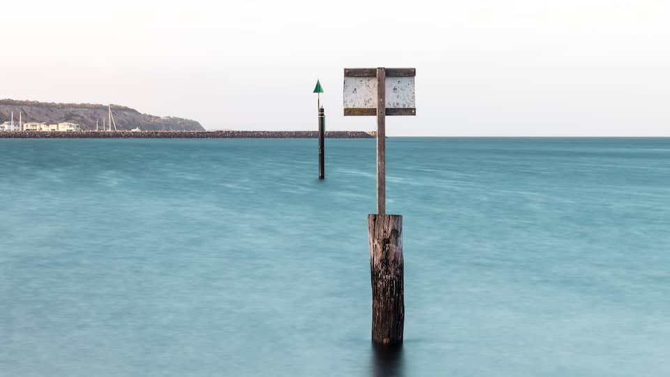 A wooden post with a sign in the water, a distant shoreline, and a clear sky.