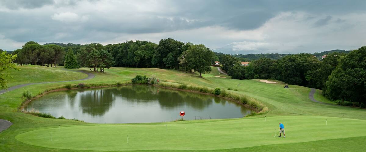 Golfista junto ao lago ao fundo a dar a tacada na bola num dia muito nublado
