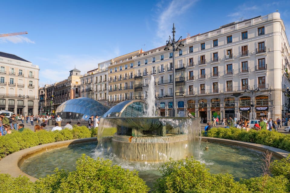 A public square with a fountain, surrounded by lush greenery and historic buildings.