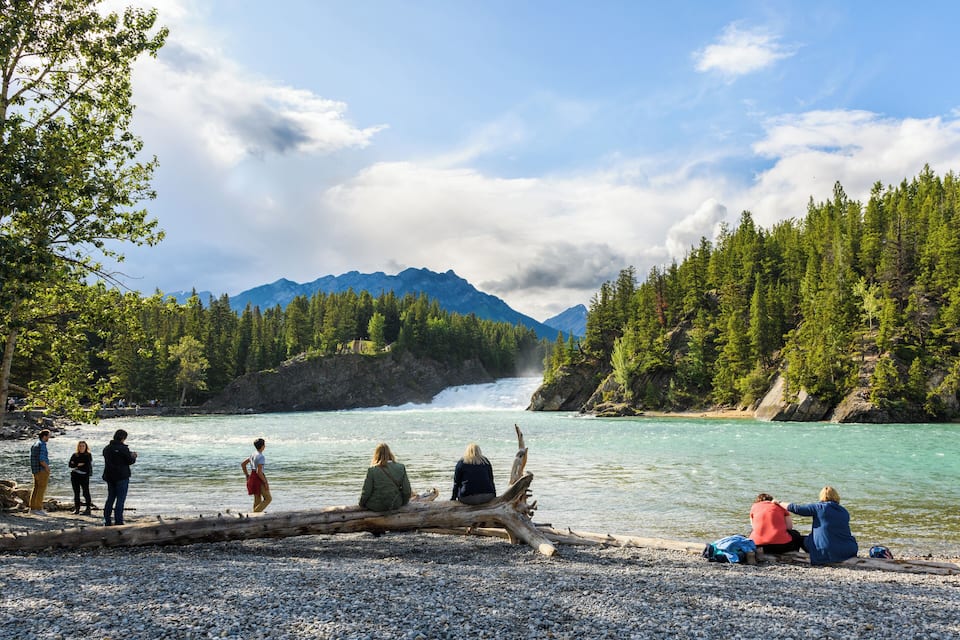 Eine Gruppe von Menschen sitzt an einem Baumstamm an einem Fluss, im Hintergrund Berge.