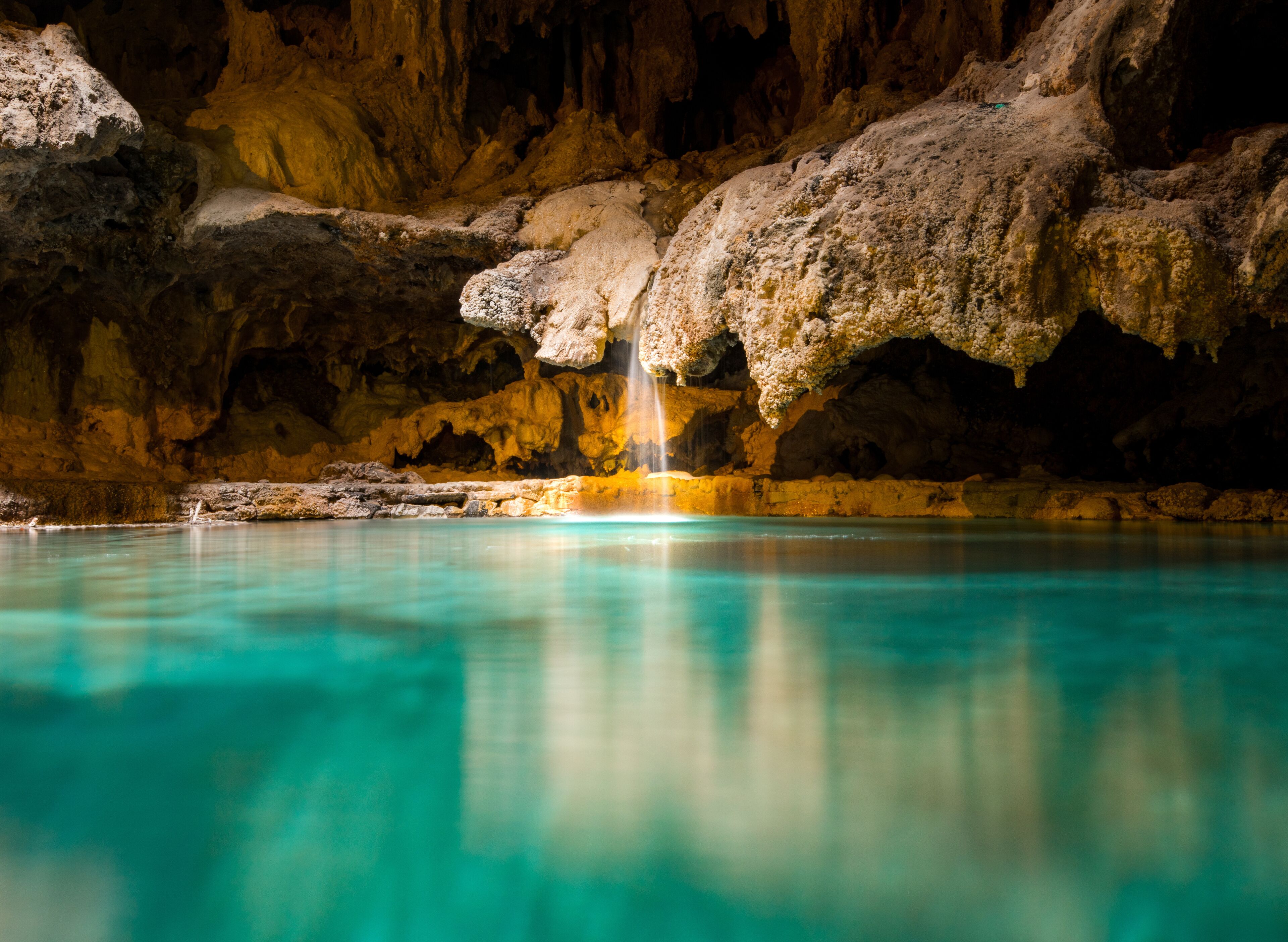 A small picturesque waterfall in a cave falls into a warm blue and green underground hot spring 