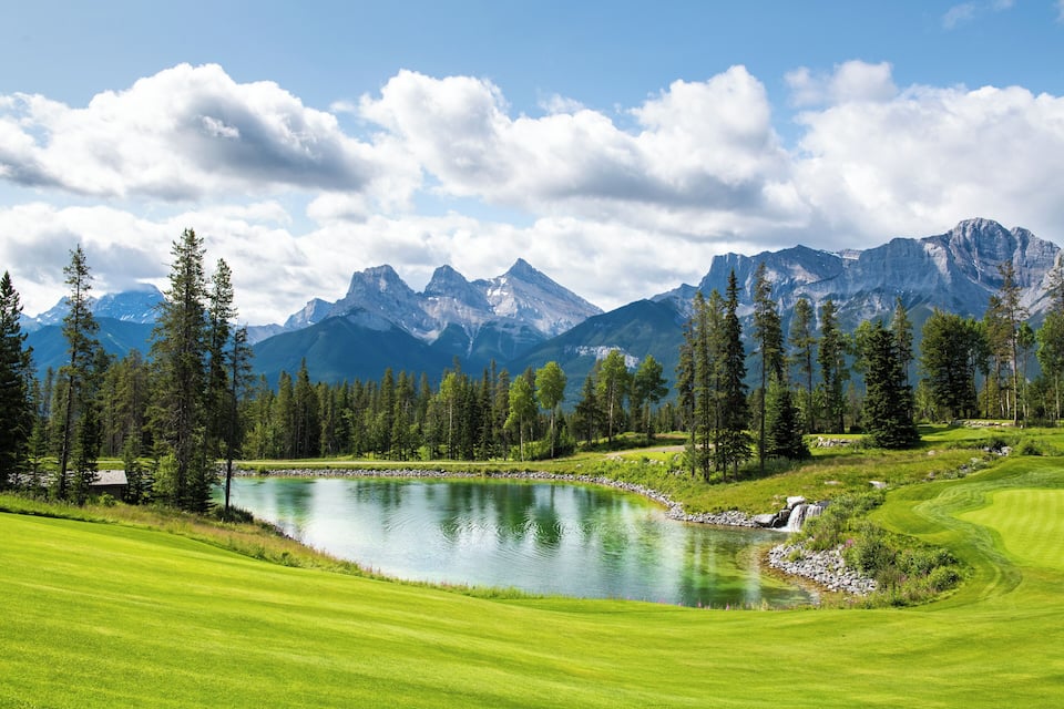 A lush green golf course with a calm lake, surrounded by tall pine trees and majestic mountains under a partly cloudy sky.