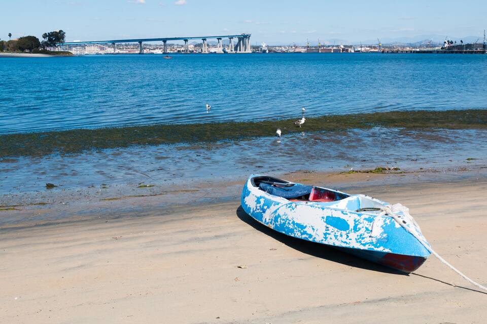 Una canoa azul y blanca atada a una playa arenosa, con un puente al fondo.