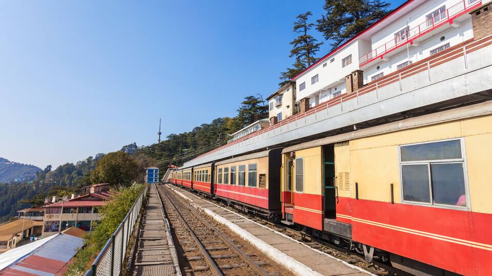A red and yellow train on a railway track with a platform and buildings in the background.
