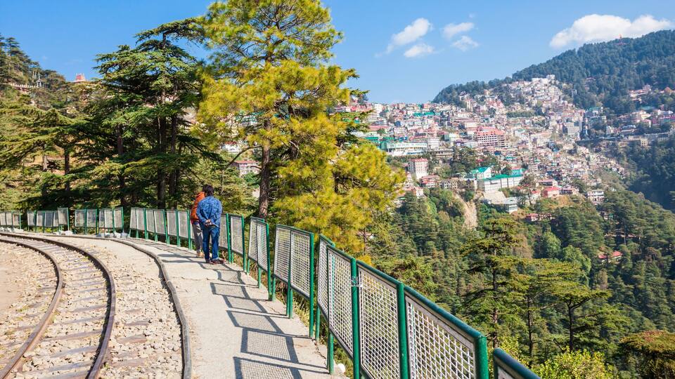 A railway track with a pedestrian walkway, surrounded by trees and a view of a hillside town.