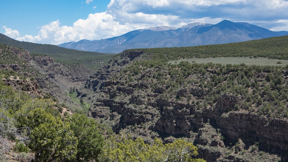 A rugged canyon landscape with green vegetation and distant mountains under a partly cloudy sky.