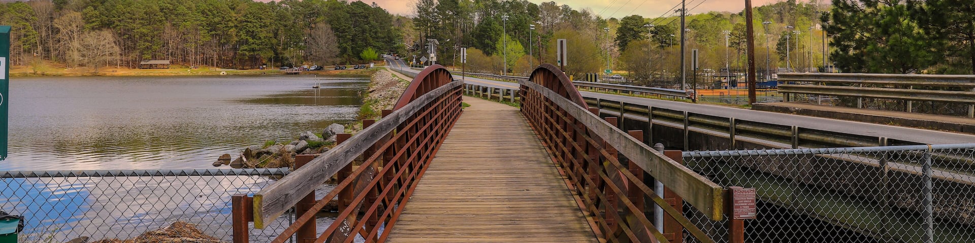 a metal and wooden rust colored bridge over a lake surrounded by lake water, green grass and lush green trees with powerful clouds and red sky at sunset at Murphey Candler Park in Atlanta Georgia USA