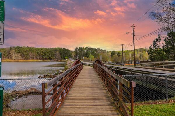 a metal and wooden rust colored bridge over a lake surrounded by lake water, green grass and lush green trees with powerful clouds and red sky at sunset at Murphey Candler Park in Atlanta Georgia USA
