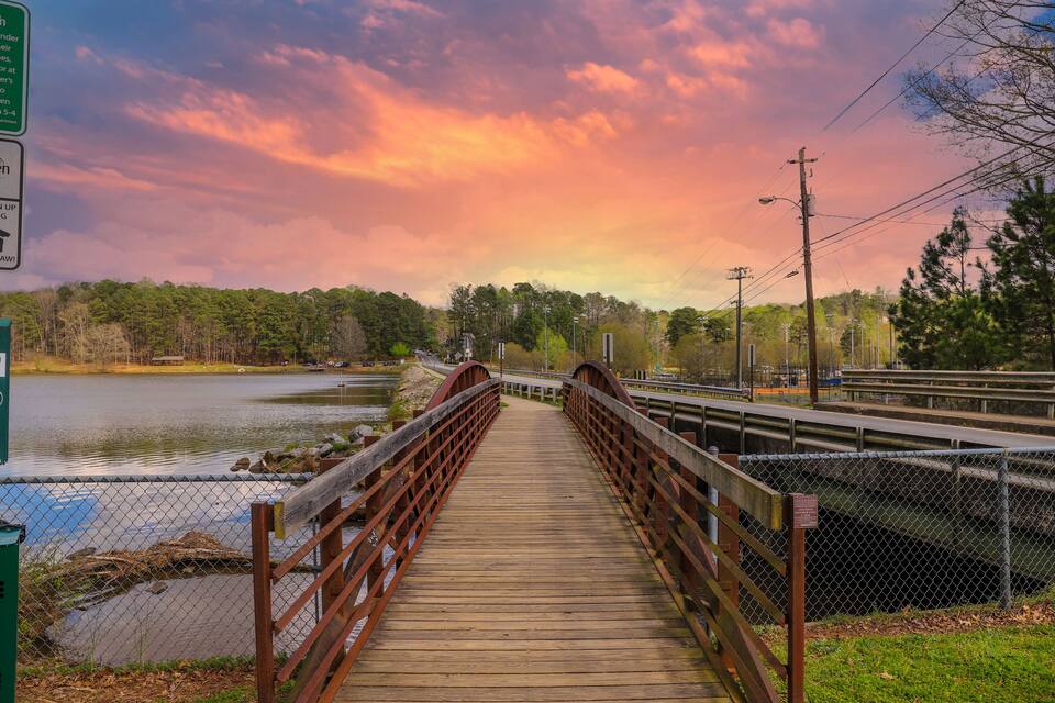 A wooden bridge over a river with a metal railing, surrounded by trees and a sunset sky.