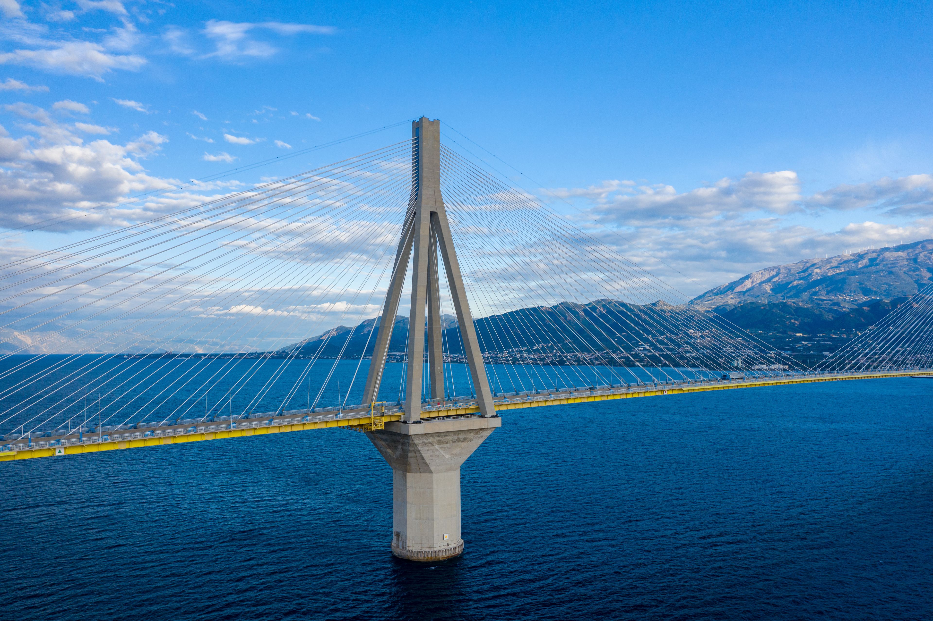 The Rion-Antirion Bridge near Patras in western Greece rises above deep blue water, with striking suspension cables and distant mountains under a vibrant sky. The structure's geometric lines stand out