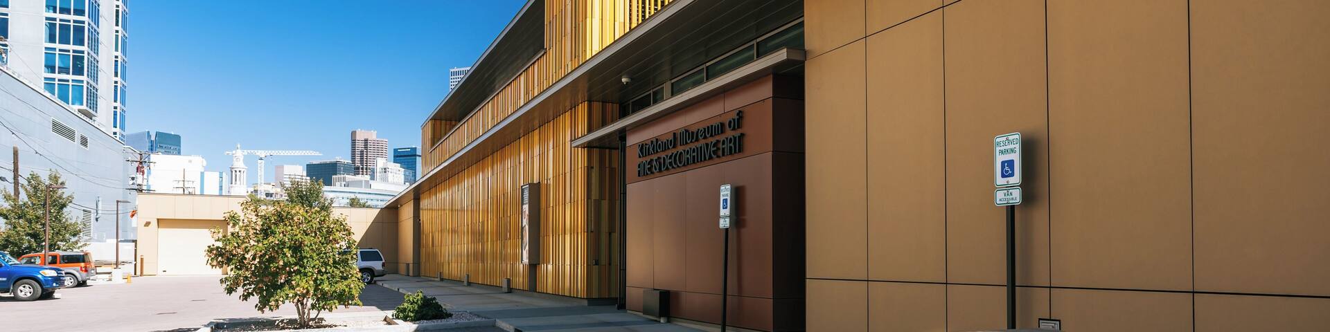 Exterior del Museo Kirkland de Arte Decorativo y Bellesas, mostrando un edificio moderno con un letrero visible, señal de estacionamiento y un pequeño árbol al frente.