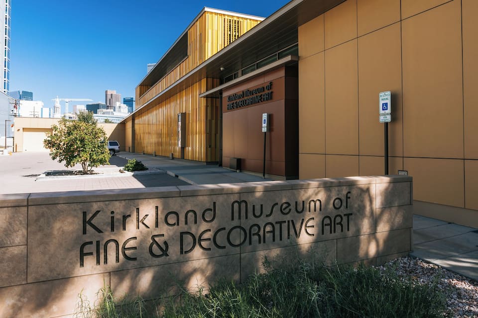De buitenkant van het Kirkland Museum voor Schone en Toegepaste Kunsten, met een modern gebouw, een duidelijk bord, een parkeersign en een kleine boom op de voorgrond.