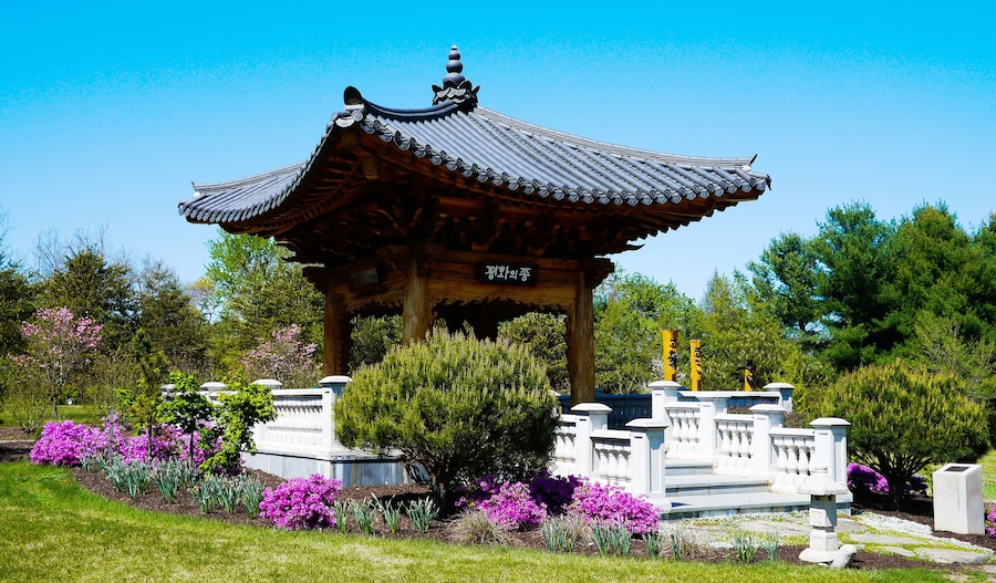 A traditional Korean pavilion with a tiled roof, surrounded by vibrant flowers and greenery.