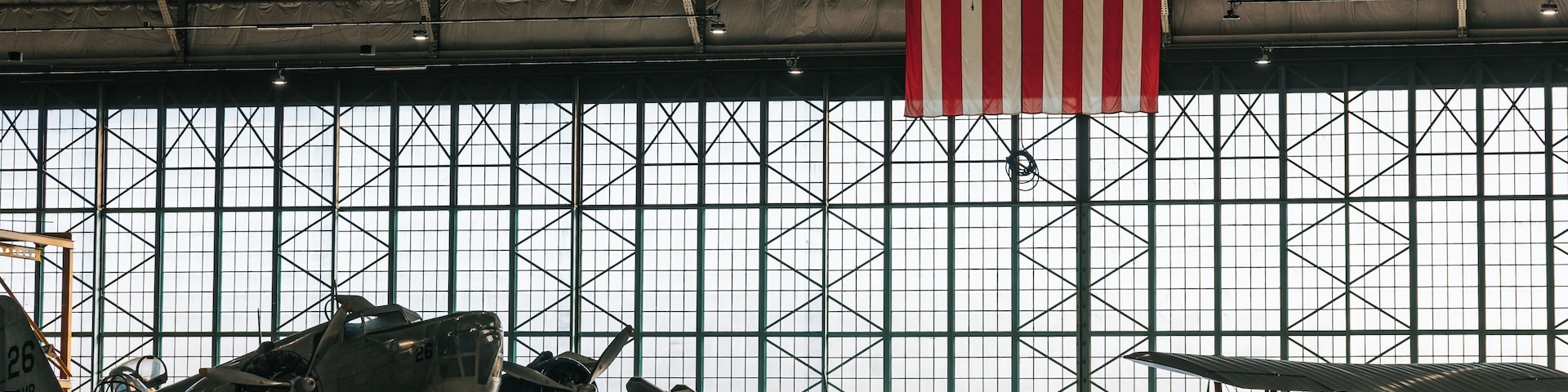 Um hangar com várias aeronaves antigas, uma bandeira americana e uma grade de janelas.