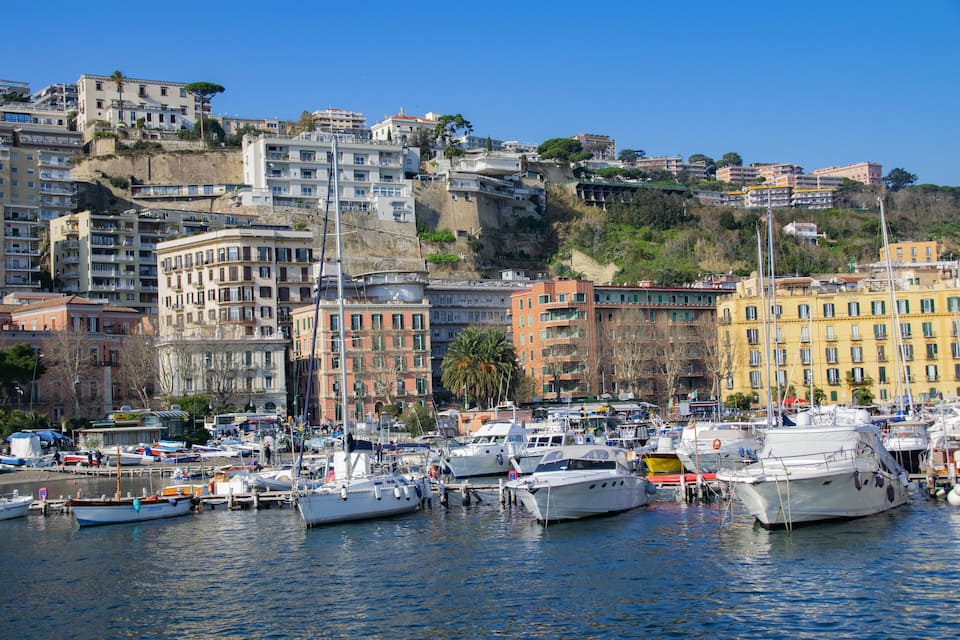 A marina with numerous boats docked, surrounded by colorful buildings and a hillside in the background.