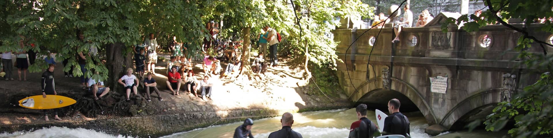 A group of people in wetsuits near a river with rapids, some holding surfboards.