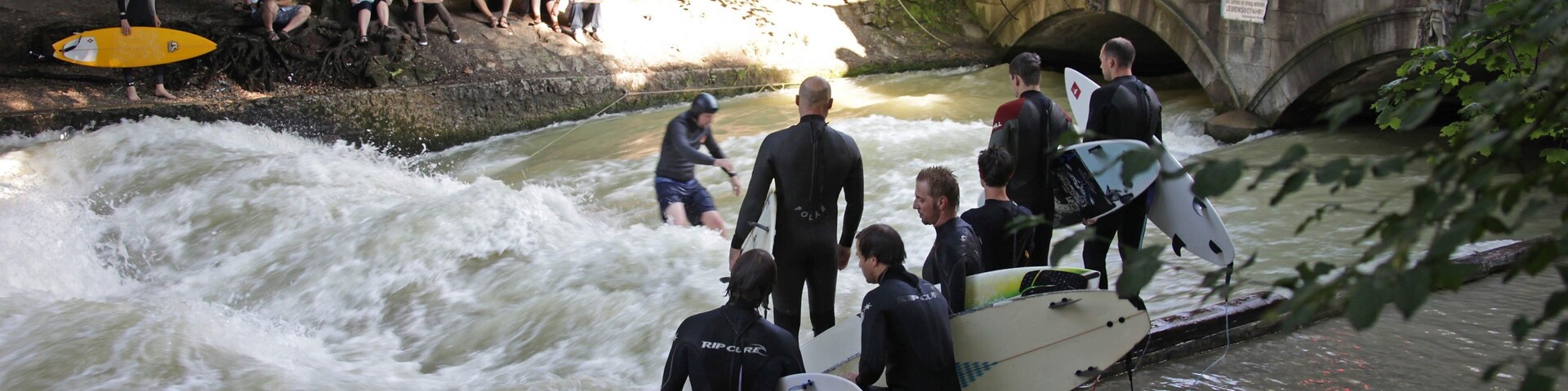 Un grupo de personas con trajes de neopreno cerca de un río con rápidos, algunas sujetando tablas de surf.
